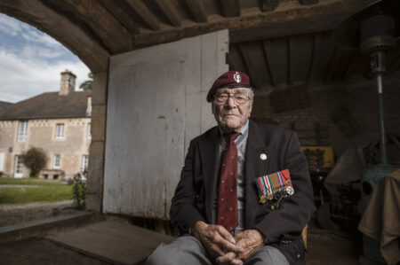 Bill Gladden sits down in a barn in Ranville, wearing military uniform including his Airborne Division beret and medals.