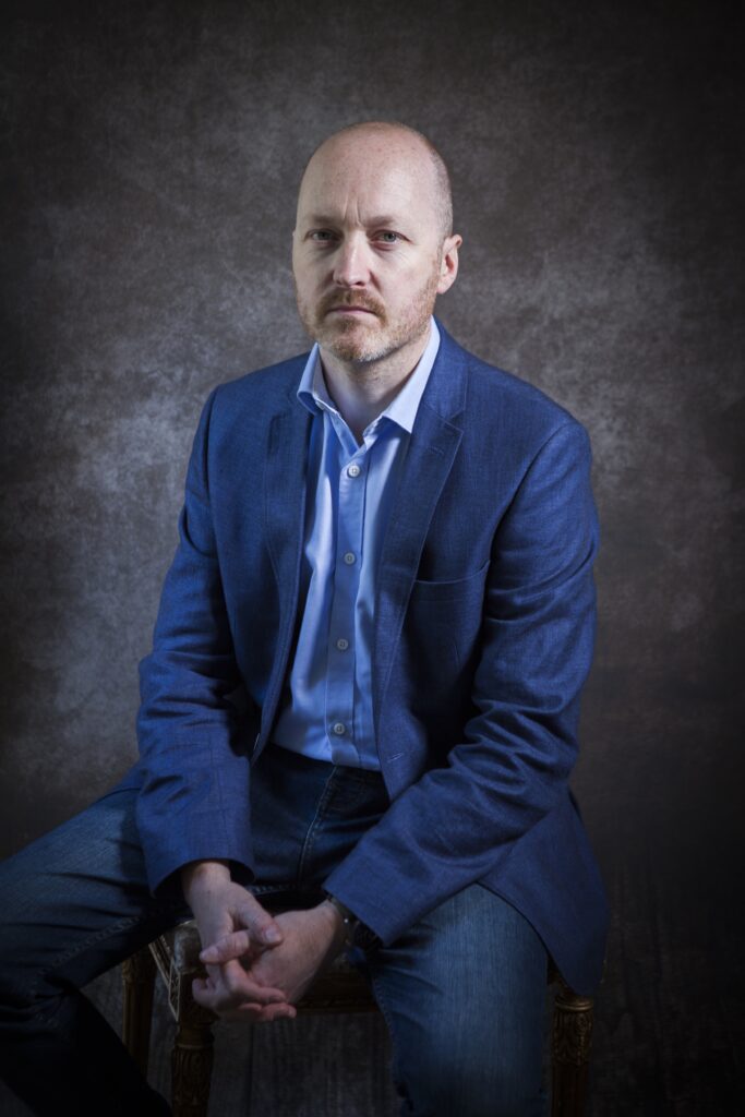 Nick Lloyd sitting against a grey background. he is wearing a blue suit, with a blue shirt and jeans.